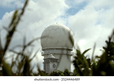 Capturing the Meteorological Radar Observatory on Sandiaojiao, uniquely shaped like a giant soccer ball, partially obscured by grass and foliage, against a cloudy sky backdrop. - Powered by Shutterstock