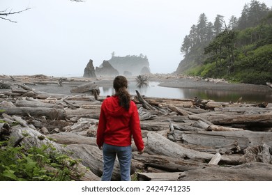 Capturing a moment of solitude, a woman clad in a red jacket stands surrounded by the vast driftwood of a foggy Pacific Northwest shoreline, reflecting on the serene and wild coastal landscape. - Powered by Shutterstock