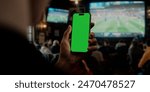 Capturing a moment in a sports bar, this image features a hand holding a smartphone with a green screen, with a lively crowd watching a soccer match in the background. Ideal for sports app promotions