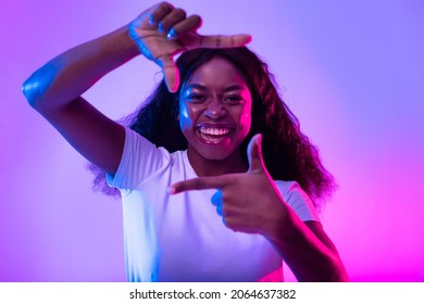 Capturing moments. Happy black woman making picture frame with fingers, looking at camera and smiling for photo in neon light. African American lady showing photograph gesture