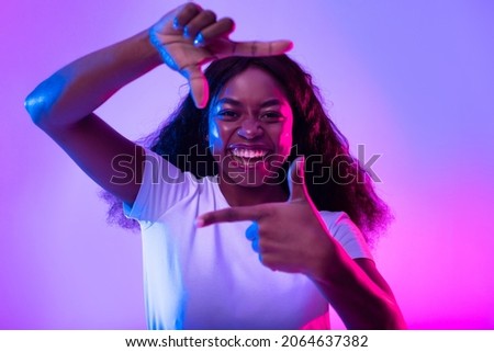 Capturing moments. Happy black woman making picture frame with fingers, looking at camera and smiling for photo in neon light. African American lady showing photograph gesture
