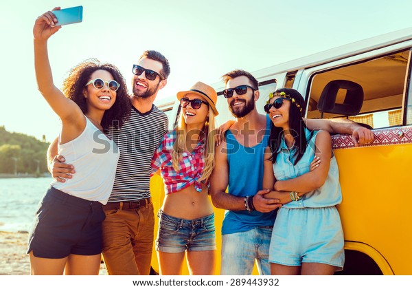 Capturing summer fun. Group of happy young people\
bonding to each other and making selfie while standing near their\
retro mini van