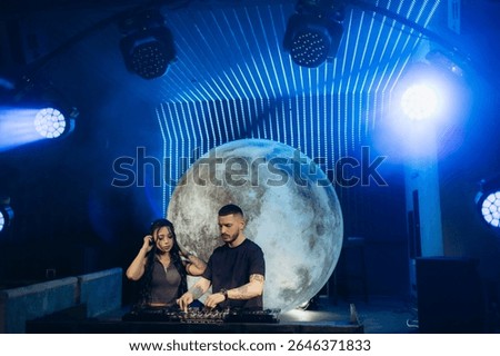 Capturing two DJs operating a turntable in a vivid nightclub scene illuminated by special moon-themed lighting, enhancing the atmospheric mood.