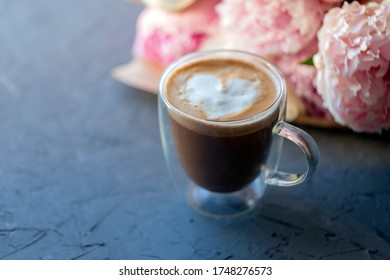 Capuccino In Double Glass Cup, Milk Foam Heart On Top, Peony Flowers Defocused On The Background.