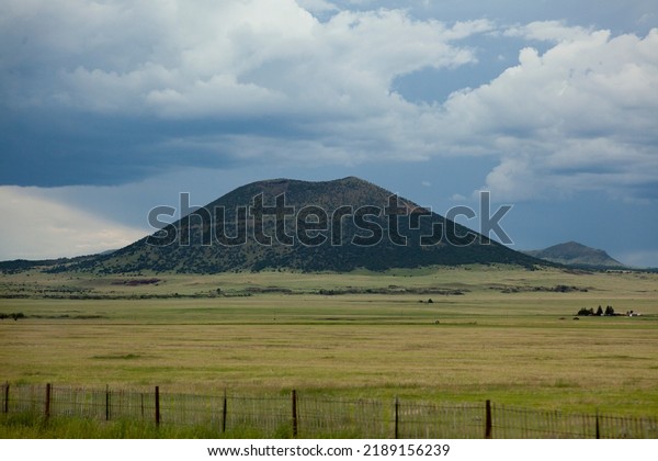 Capulin Volcano in New Mexico on a cloudy summer\
day with blue skies and green grass in this dramatic view from a\
road trip.