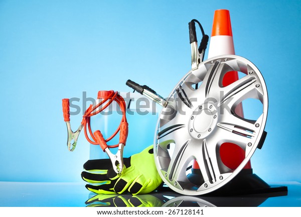 Car accessories - car hubcap with jump start
cables in front of traffic cone with washer fluid and a pair of
green gloves on blue
background