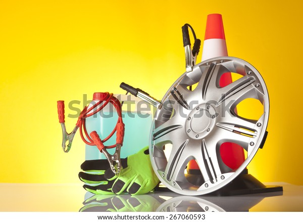 Car accessories - jump start cables
tangled on washer liquid with a pair of green gloves next to alloy
wheel in front of traffic cone on yellow
background