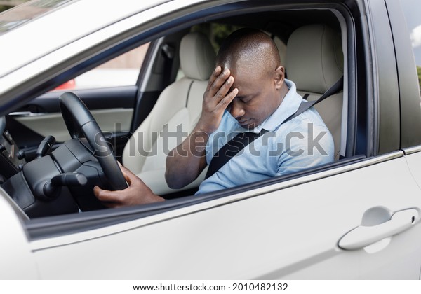 Car accident concept. Upset black man driver sitting\
at front seat and touching his head, having automobile crash.\
African american guy getting late for flight, got stuck in traffic,\
side view