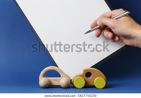 Car Accident Insurance Concept Drivers hand\
with pen white sheet of paper wooden toy cars with symbolizing car\
accident on blue\
background.