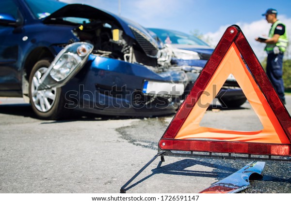 Car accident. Road disaster. Vehicle\
collision. Warning triangle for traffic and damaged cars in the\
middle of an intersection. In background police officer writing an\
accident report. Car\
insurance.