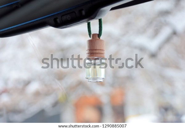 Car air perfume freshener inside the car with blurred\
winter background. Little glass bottle with wooden lid and yellow\
aromatic liquid automobile freshener on a green rope and car mirror\
