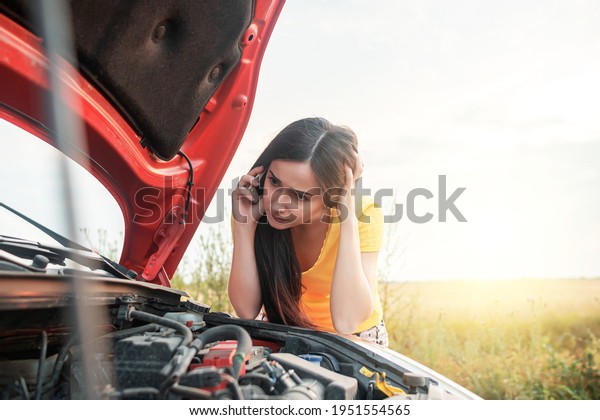 Car breakdown. Young woman\
calls on the phone while standing by a broken car with an open\
hood.
