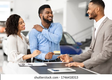 Car Buyers. Joyful Black Couple Giving Credit Card To Auto Seller In Vehicle Dealership Office. Selective Focus