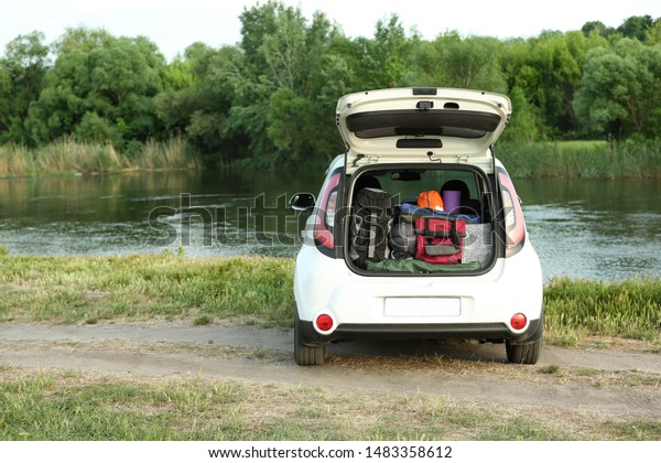 Car with camping equipment in trunk on riverbank.\
Space for text