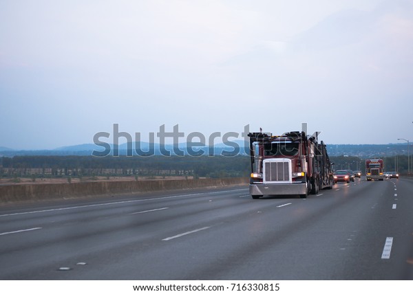 A car carrier on a large classic American big rig\
semi truck with a two-tier trailer for the transport of cars goes\
on a multi-lane highway in the evening twilight towards the loading\
point