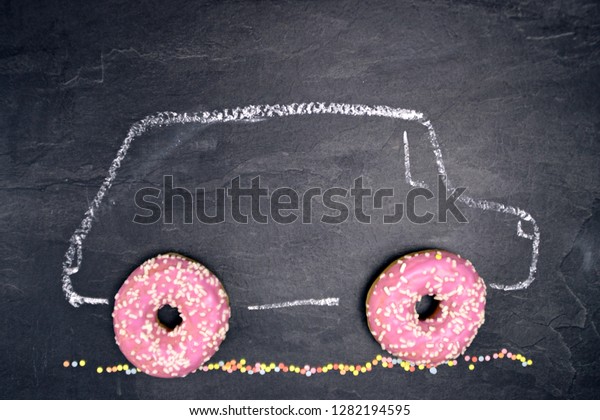 A car with chalk painted on a dark stone
surface with pink donuts with white sprinkles as wheels - Abstract
concept with donuts as wheels of a
car