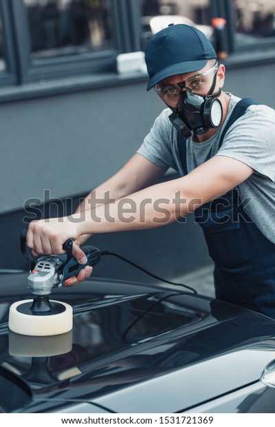 car cleaner in\
respirator and protective glasses looking at camera while polishing\
car with polish machine
