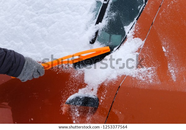 Car covered with snow.\
Brush in mans hand. Man in gray gloves is brushing orange car from\
snow.