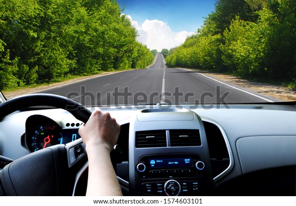 Car\
dashboard with driver\'s hand on the black steering wheel and road\
with green trees against blue sky with\
clouds