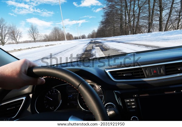 Car dashboard with driver\'s hand on the black\
steering wheel with winter countryside road with trees against a\
blue sky with clouds