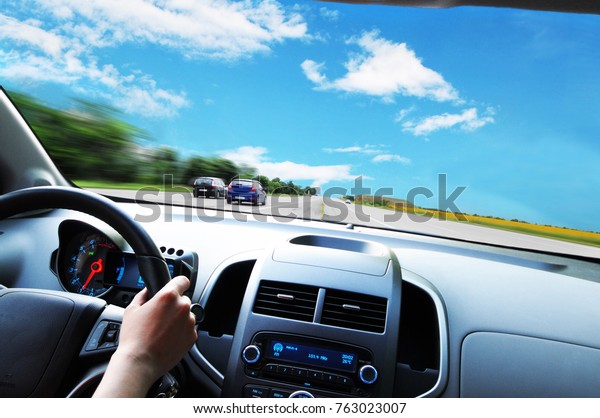 Car dashboard with\
driver\'s hand on the black steering wheel against asphalt road and\
blue sky with clouds