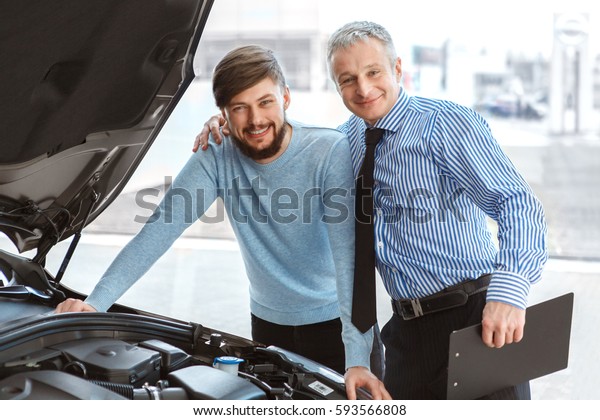 Car dealer and his client. Cheerful mature car dealer\
embracing his male client smiling happily to the camera posing near\
a new car with open hood at the dealership agreement communication\
service job