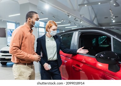 Car Dealer In Medical Mask Pointing With Hand At Auto Near African American Man