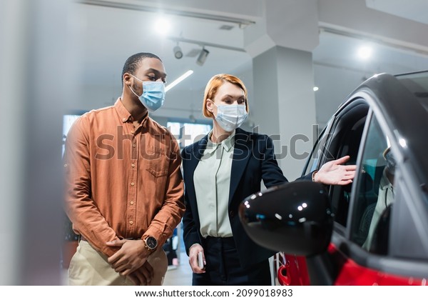 car dealer in medical mask pointing at car near\
african american customer