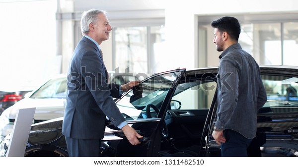 Car dealer\
opening a car\'s door to a\
customer