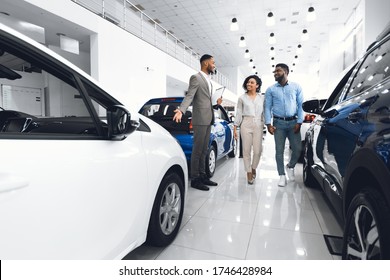 Car Dealership. Cheerful Manager Guy Showing Automobile To Customers In Auto Dealership Store. Copy Space