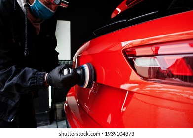 Car detailing concept. Man in face mask with orbital polisher in repair shop polishing orange suv car.