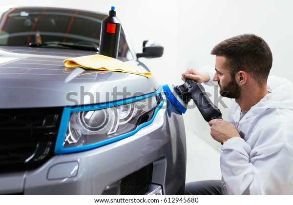 Car detailing - Hands with orbital polisher in\
auto repair shop. Front lights protected with isolation blue tape.\
Selective focus.