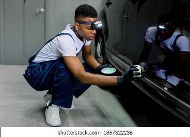 Car detailing and polishing concept. Young African man worker, wearing uniform and gloves, holds the sponge in hand and polishes the car body with solid wax. Selective focus.