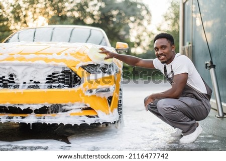 Car detailing wash at outdoors car wash service. Handsome African American man in white t-shirt and overalls washing car radiator grille and hood of his new yellow sport car with foam and sponge