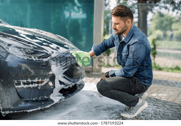 Car detailing wash at\
outdoors car wash service. Handsome bearded man in casual wear,\
washing car radiator grille and hood of his new electric car with\
foam and sponge.