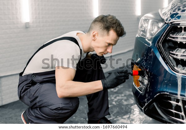 Car detailing\
wash. Professional car service Caucasian male worker in gray\
overalls and black rubber gloves, doing detailing wash and cleaning\
the car with special black\
brush