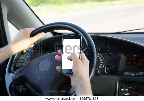 Car driver presses the blank screen of smartphone
with his thumb while driving along the road. Copy space in display
frame. Empty touchscreen in men's right hand. Left hand holding a
steering wheel.