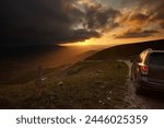 The car drives along a dirt road in the background of a landscape in the mountains and a bright sunset sky. Rear view with bumper
