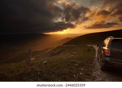 The car drives along a dirt road in the background of a landscape in the mountains and a bright sunset sky. Rear view with bumper
