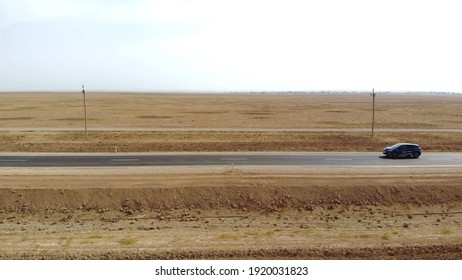 A Car Drives By On An Empty Road. The Drone Follows The Car On A Narrow Asphalt Road. The Car Drives Through The Desert. Side View Of A Passing Car