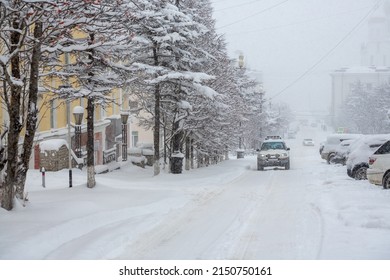 A Car Drives Down A Snow-covered City Street During A Heavy Snowfall. A Lot Of Snow On The Roadway, Sidewalks And Trees. Snowdrifts On The Sides Of The Road. Snowstorm In The City. Magadan, Russia.