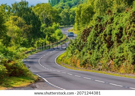 A car drives on a road. A narrow, winding road in Scotland along Loch Ness. Trees and bushes next to the road in summer in sunshine. Traffic signs and guard rails