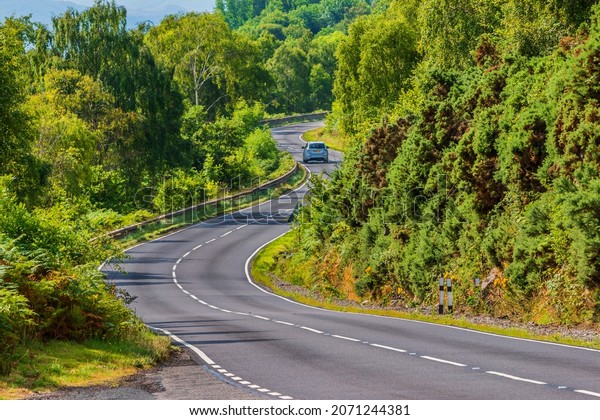 A car drives on a road. A\
narrow, winding road in Scotland along Loch Ness. Trees and bushes\
next to the road in summer in sunshine. Traffic signs and guard\
rails