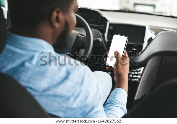 Car Driving App. African American Man Using Phone\
With Empty Screen Sitting In Auto Vehicle. Selective Focus, Mockup,\
Back View