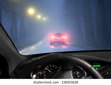 Car Driving In A Dark Avenue In Thick Fog, Seen Through Windscreen Of Other Vehicle