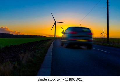 Car Driving Down Country Road Towards Wind Turbines In A Field In The UK At Sunset Or Sunrise On  A Clear Winter Day