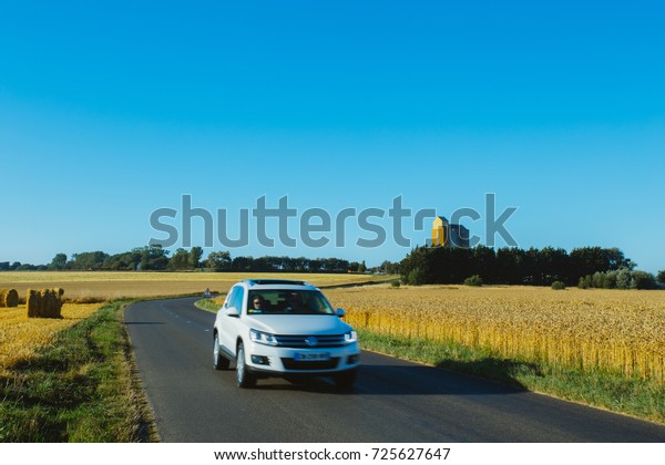 Car driving on asphalt country road through\
yellow wheat field. Country landscape on a sunny summer day in\
France. Environment friendly farming and agriculture, harvest\
season, transportation\
concept