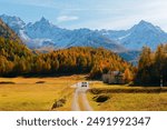 A car driving on a country road in Bernina Pass on a brisk fall day, with golden larch forests and snowy alpine mountains under blue sky in background, in canton of Grisons (Graubünden), Switzerland