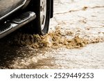 A car is driving through a muddy road with water splashing out from the tires. The scene is chaotic and messy, with the water covering the ground and the car