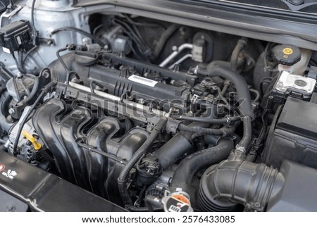 A car engine bay with various components including the intake manifold, hoses, wiring, and fluid reservoirs. The engine appears to be a modern internal combustion engine.